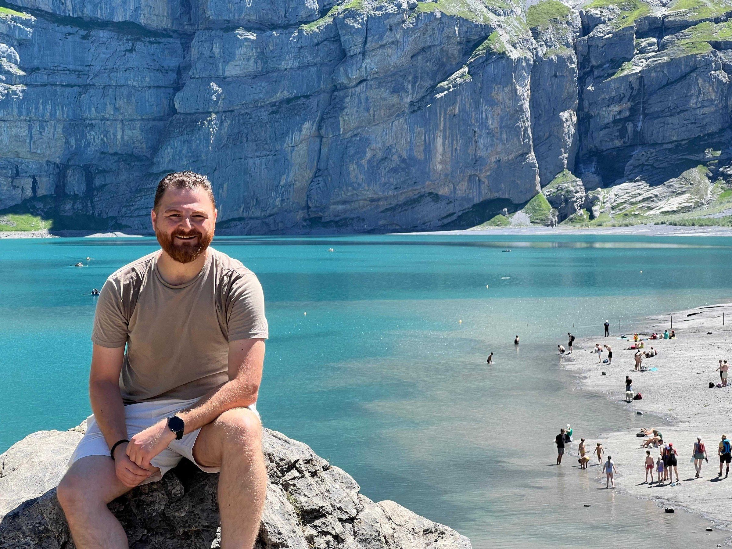 Muhannad Satouf smiling, sitting on a rock above a turquoise mountain lake
